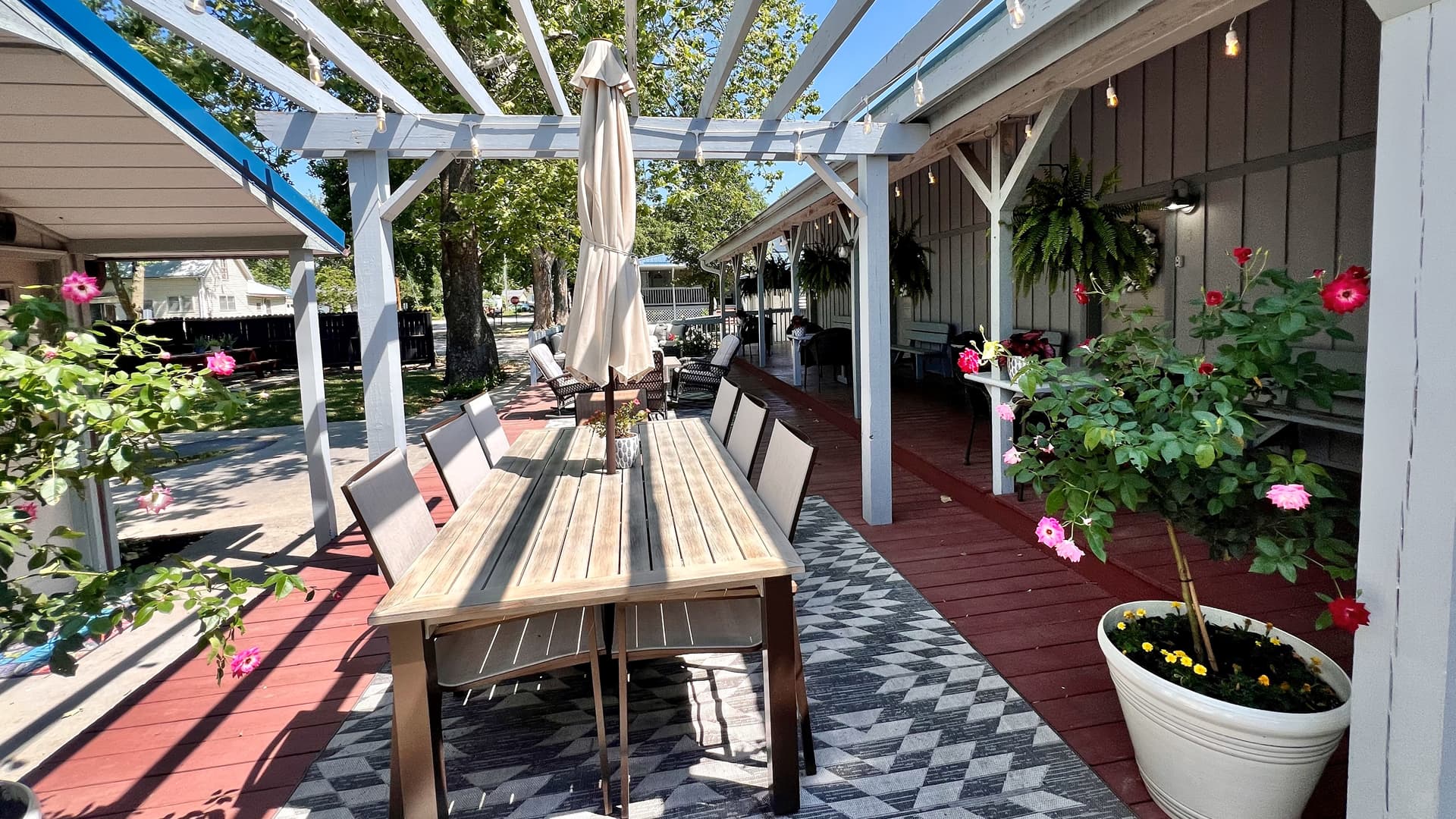 arbor trellis with pink flowers, hammock chairs and metal patio furniture on a cement patio between two white wood buildings with a green grass yard in the foreground