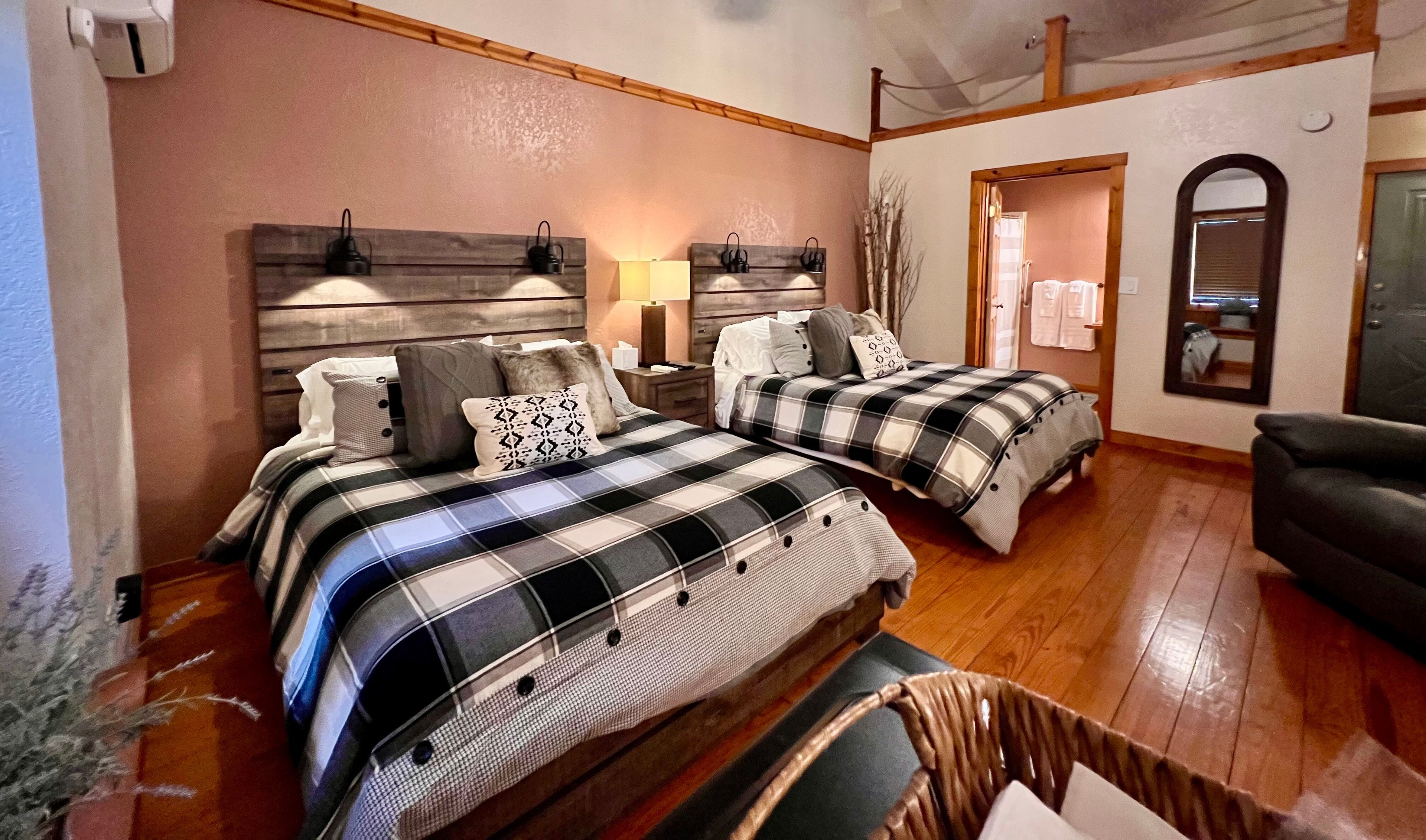 Two lower queen beds side by side topped with black & white buffalo plaid duvets and decorative throw pillows, lighted rustic headboards, doorway leading to bathroom