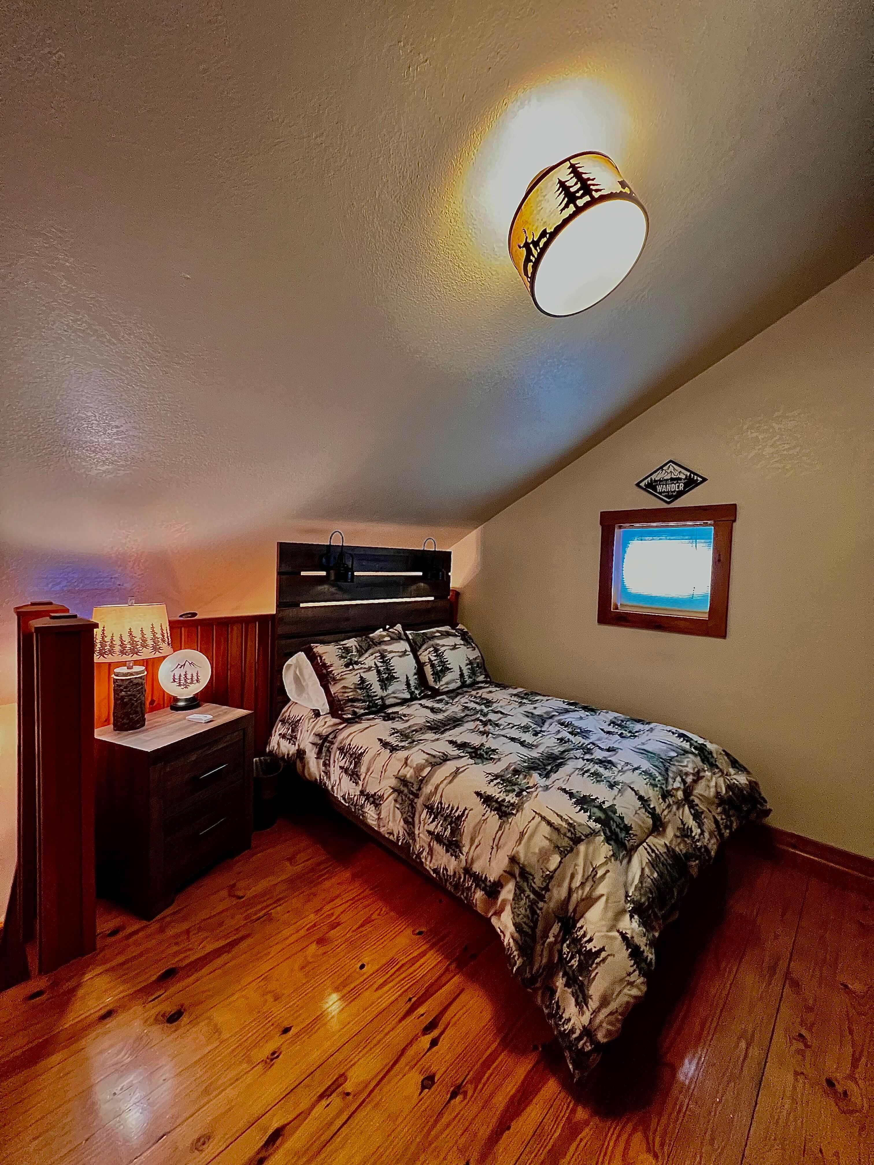 Loft bedroom with full-size bed featuring bedding with pine tree print, nightstand with rustic table lamp, beautifully finished wood floors, and bean bag chair (not shown)