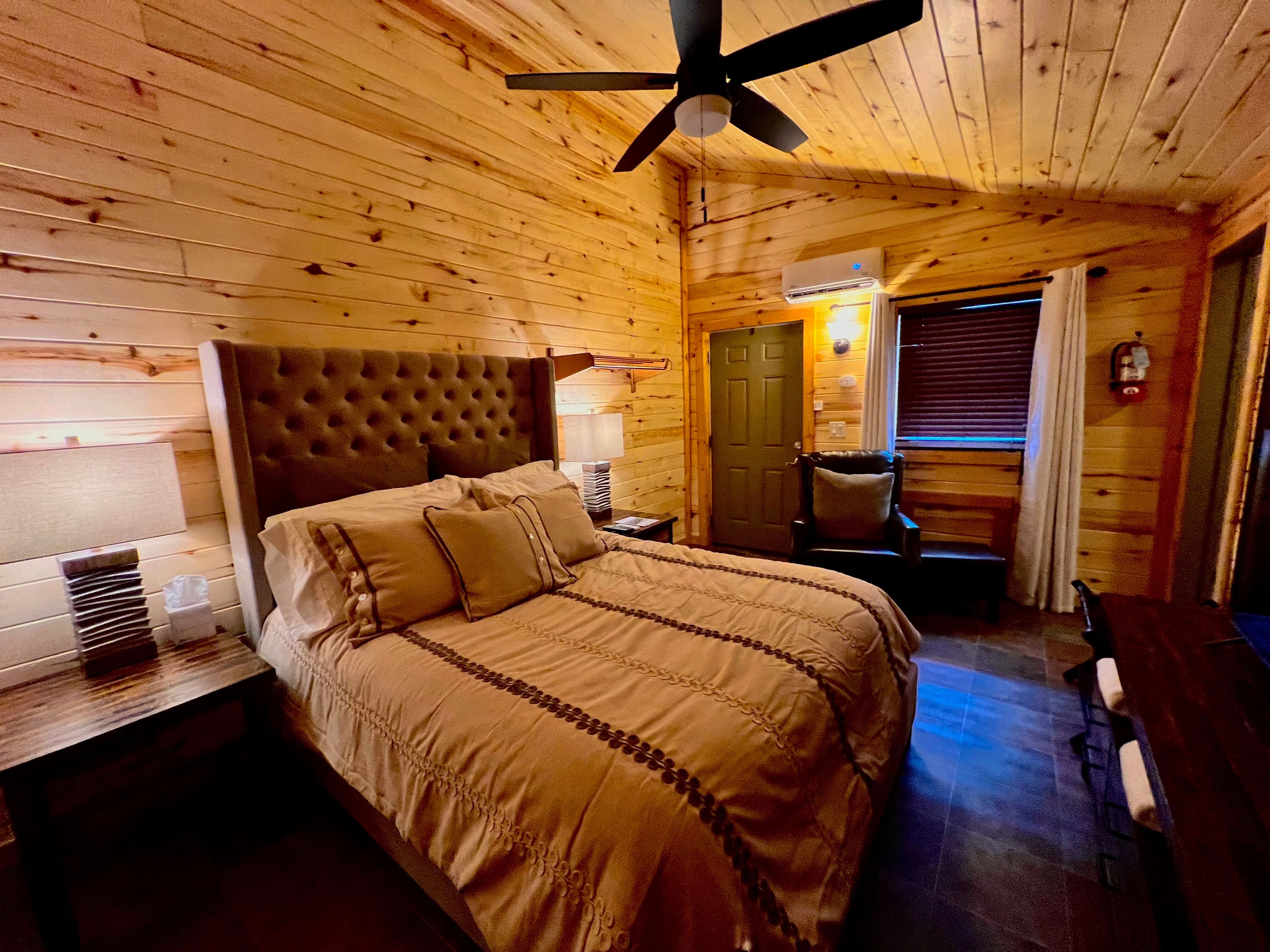 Warmly lit bedroom with queen bed featuring button-tufted upholstered headboard, natural pine walls and ceiling, ceiling fan