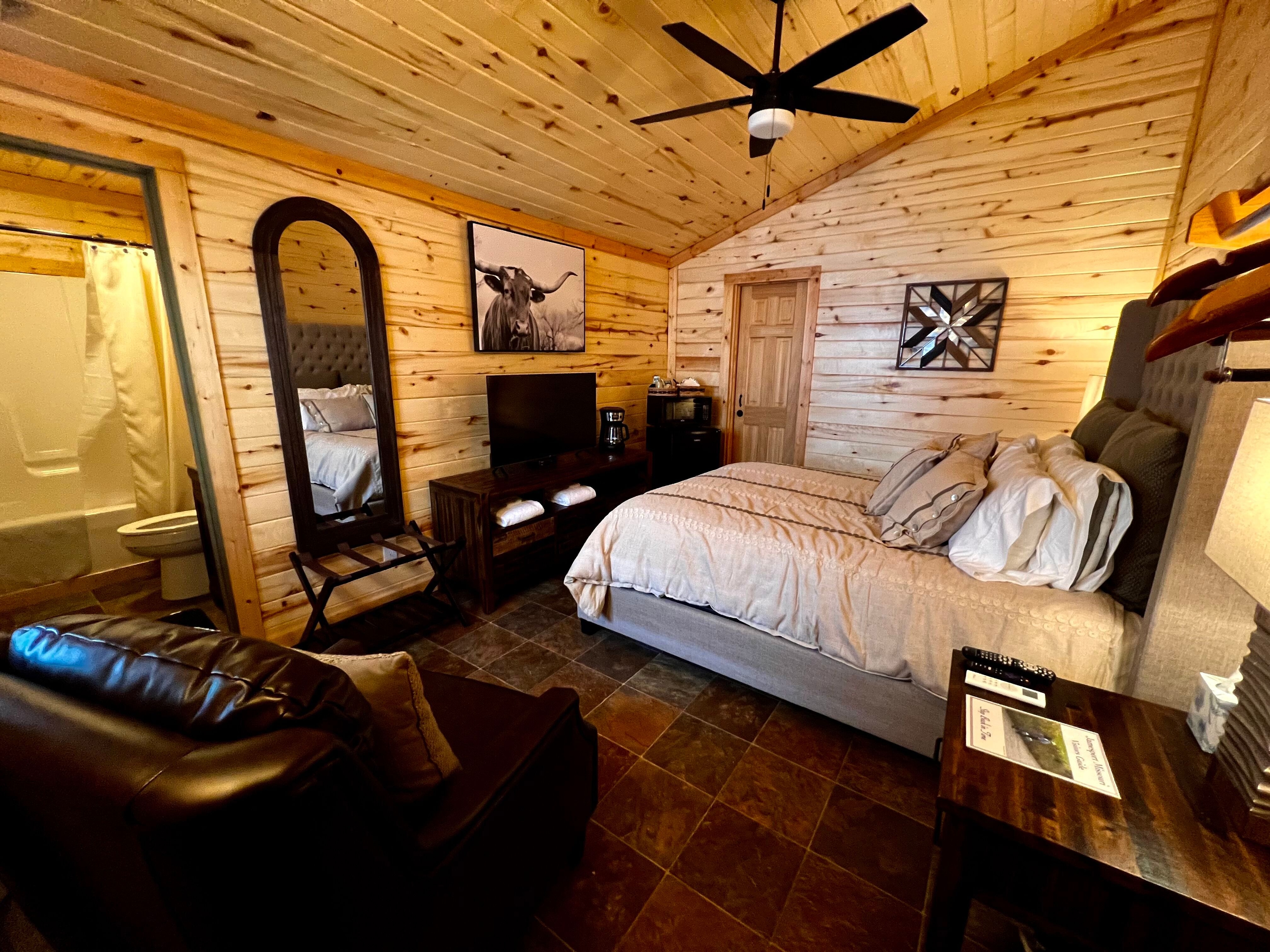 Sleek rustic room with queen bed, nightstands, natural pine walls, overstuffed brown leather chair, console with flat-screen TV, doorway leading to bathroom