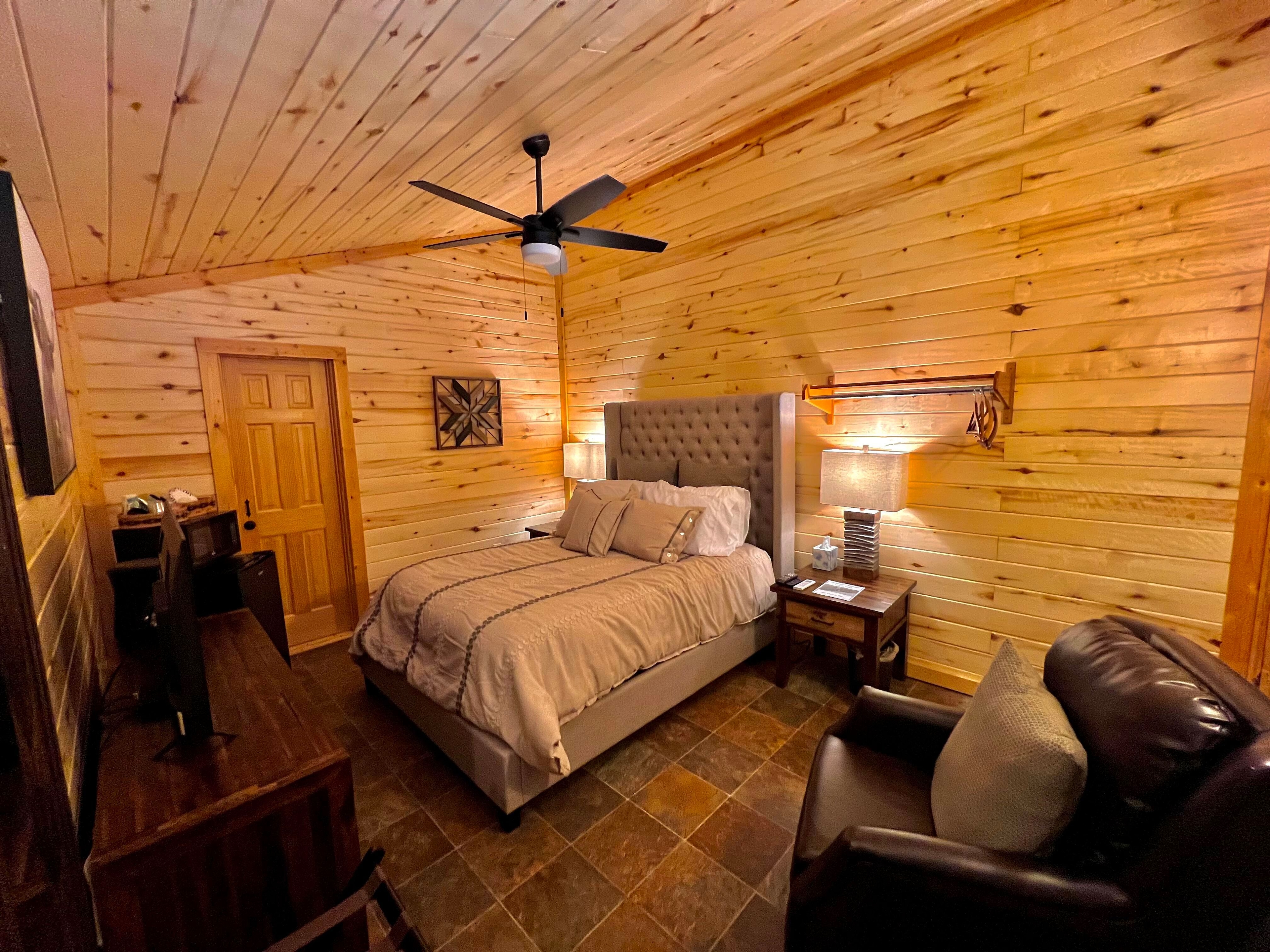 Modern rustic bedroom with natural wood walls and ceiling, ceiling fan, upholstered headboard, overstuffed brown leather chair, and TV console