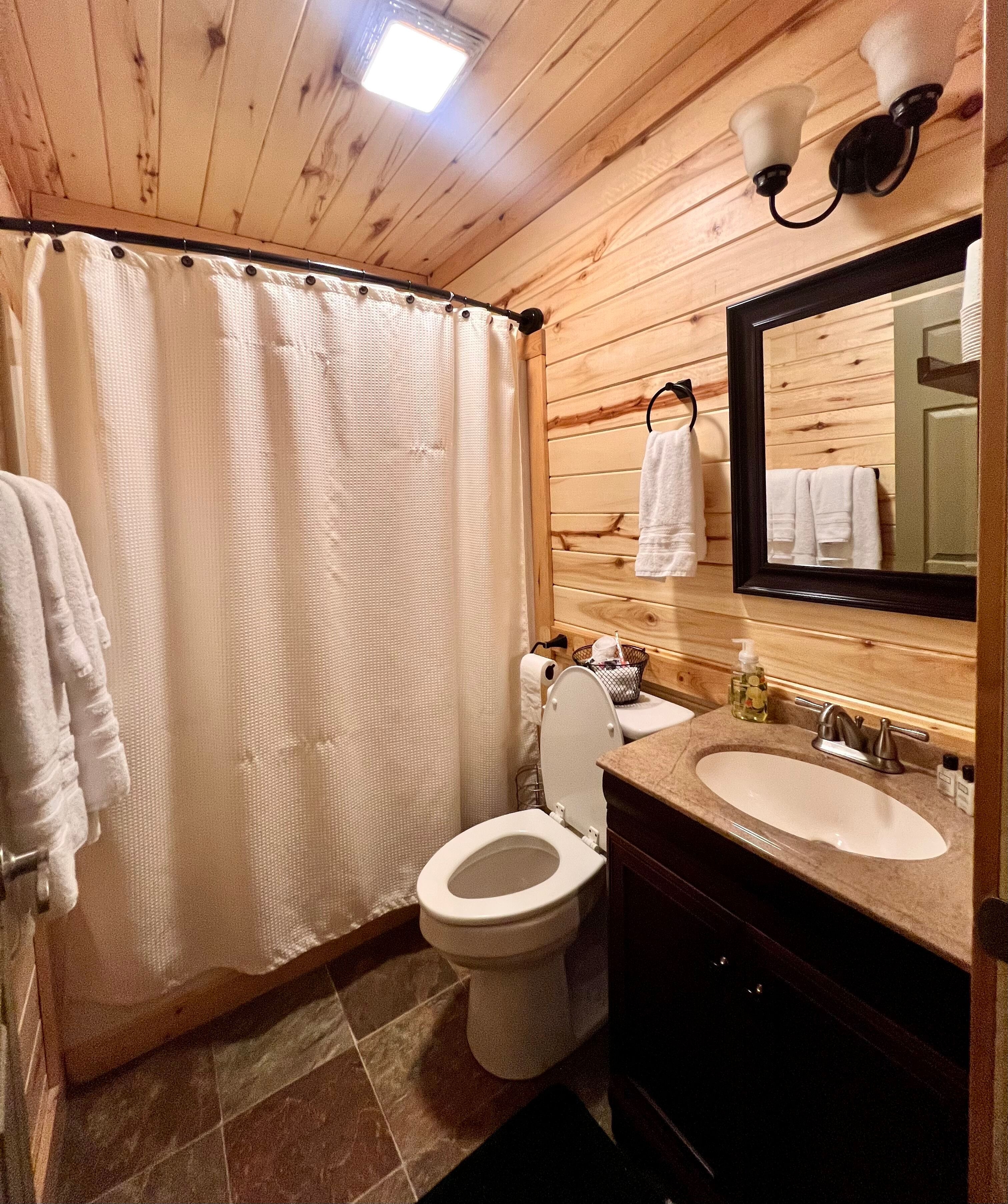 Bathroom with vanity sink and mirror, toilet, shower, and plush white towels hanging on natural wood walls