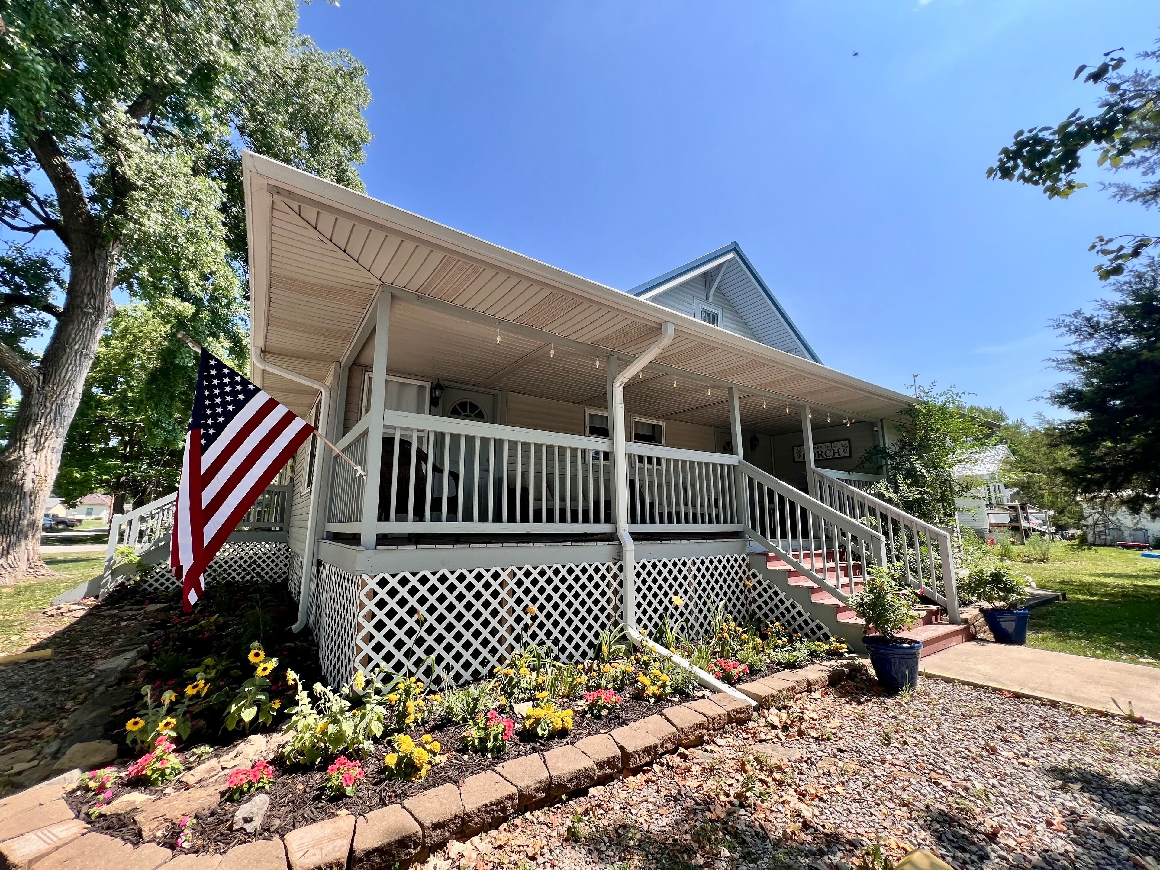 Two-story house with front steps leading to large front porch, landscaping with flowers, blue skies, and an American flag on a pole waving in the wind