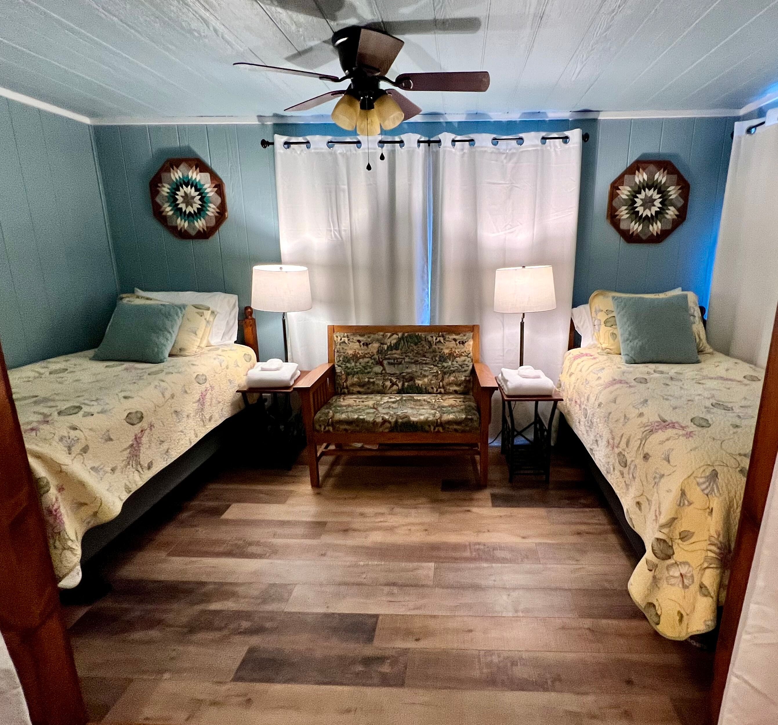 Peaceful bedroom with blue walls, two single beds separated by an upholstered bench and matching nightstands with table lamps, and large window with white drapes, wide plank wood floor, and ceiling fan